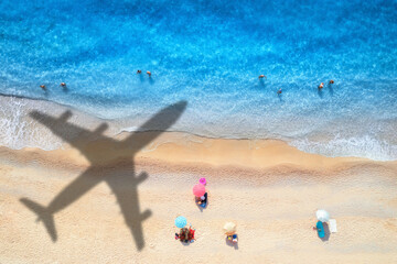 Aerial view of blue sea, airplane shadow, sandy beach with umbrellas at sunset in summer. Tropical landscape with blue water, people, shadow of flying plane. Travel and vacation in islands. Top view