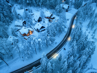 Aerial view of fairy village in snow, road, forest, houses with illumination, street lights at winter night. Top view of alpine countryside, snowy pine trees at dusk. Kranjska Gora, Slovenia
