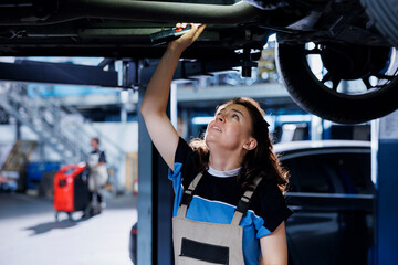 Meticulous mechanic working on suspended car in garage, checking motor during routine maintenance. Auto repair shop employee underneath vehicle, inspecting parts using work light