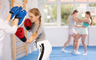 Concentrated sporty teen girl in boxing gloves practicing self-defense techniques in gym, throwing punches on focus mitts in hands of instructor..
