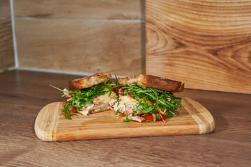 Large satisfied sandwich splited in two consist from slices of roasted white bread, pork ham, dutch cheese, sunny side up egg and rocket salad on wooden board as a main course for lunch or dinner.