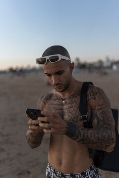 Chico joven tatuado y musculoso sin camiseta tomando fotos con el smartphone en la playa