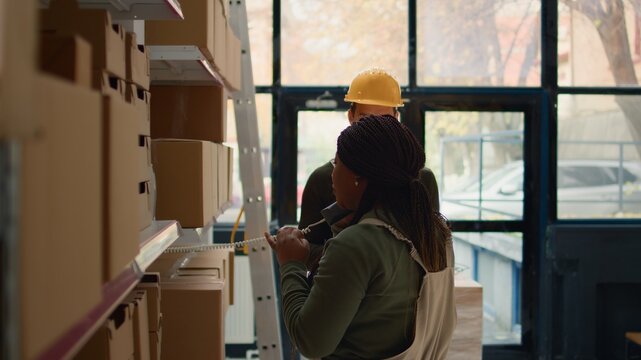 African american supervisor and intern preparing warehouse orders for delivery, using tablet to verify shipping informations, receiving phone call while scanning stock labels in distribution center