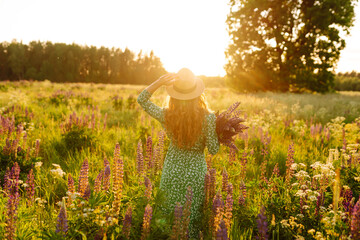 A young woman in a stylish green dress and hat enjoys the sunset in a field with lupins. A beautiful woman with a bouquet of lupins, collects flowers, inhales fresh floral air. 