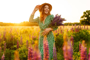 A young woman in a stylish green dress and hat enjoys the sunset in a field with lupins. A beautiful woman with a bouquet of lupins, collects flowers, inhales fresh floral air.  - obrazy, fototapety, plakaty