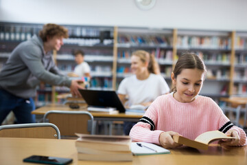 Fototapeta premium Portrait of a ten-year-old schoolgirl sitting at a desk in the school library and reading an interesting book