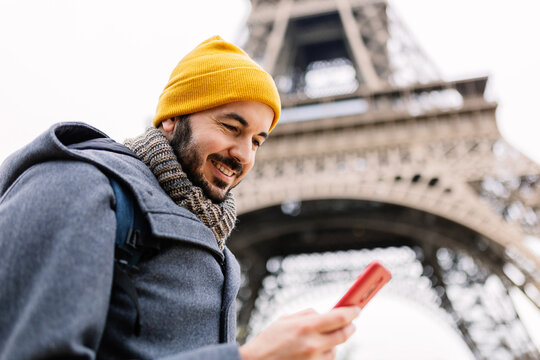 Happy young adult tourist man using mobile phone close to Eiffel Tower in Paris. Communication and travel concept.