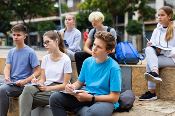 Attentive students write lesson in a notebook sitting on a stone street parapet - obrazy, fototapety, plakaty
