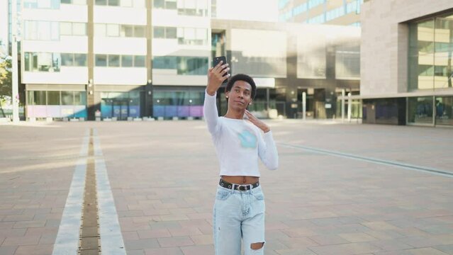 Cute non-binary person taking a selfie with their smartphone and sharing it on their social networks. Cheerful black person taking a selfie with the mobile phone having fun in the city outdoors