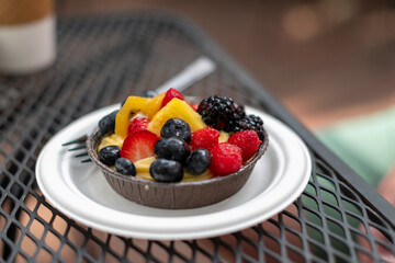 A fruit filled custard tarte. The topping has raspberries, blueberries, strawberries, and kiwi on an almond crust. A small fork in the dessert is lying on a white plate on a metal table at a cafe.