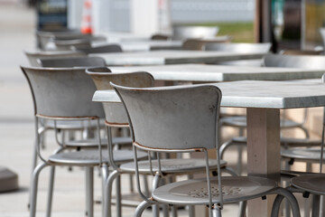 A row of multiple empty grey metal square tables and chairs outside on the sidewalk of a cafe or bakery. The chairs have round backs and are empty. The ground is a grey sidewalk.
