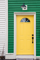 Obraz premium The exterior wall of a white and green house with a vibrant yellow colored door. There's a small semi-circular window in the solid metal door. The wall has a black light fixture over the closed door. 