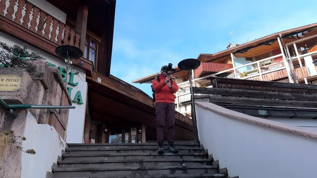 Travel Photographer With Camera On Gimbal Stabilizer Standing On Stairs, Taking Photos In Cortina d'Ampezzo, Italy. low angle, slow motion