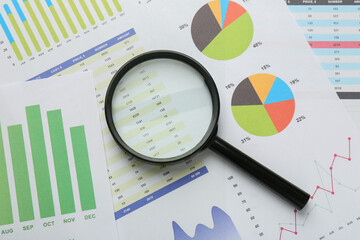 Magnifying glass on accounting documents with data and graphs, top view