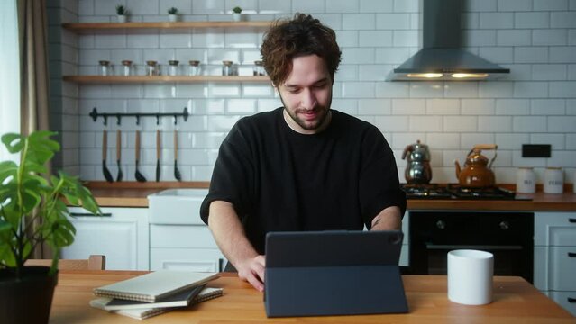 Smiling young curly hair man sitting in kitchen, using app with tablet computer, surfing on internet