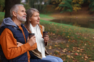 Affectionate senior couple with cups of coffee in autumn park, space for text