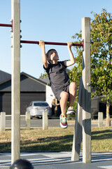 The boy plays sports on the outdoor sports field. The boy pulls himself up on the horizontal bar.