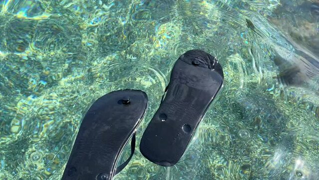 Floating black upside down flip flops in transparent turquoise sea water by the beach in Spain, fun sunny summer vacation holiday shoes, 4K shot