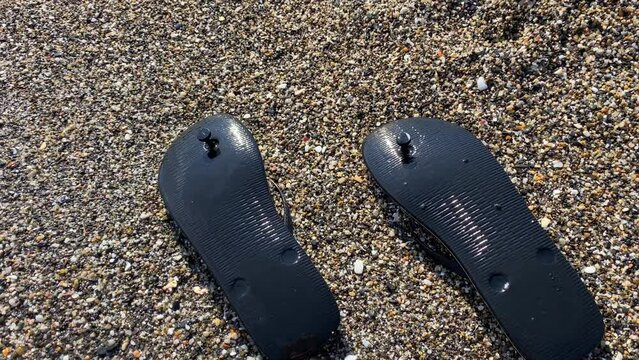 Waves hitting washed up black upside down flip flops on a beach with tiny little rocks in Spain, sunny summer vacation holiday shoes, 4K shot