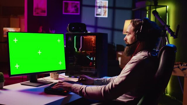 Man in dimly lit apartment playing video games on isolated screen gaming PC at computer desk, enjoying day off from work. Gamer battling enemies in online multiplayer shooter on chroma key monitor
