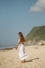Fototapeta na wymiar Happy young woman wearing beautiful white dress is walking on the beach. Summer beach vacation concept, walk at the sandy beach