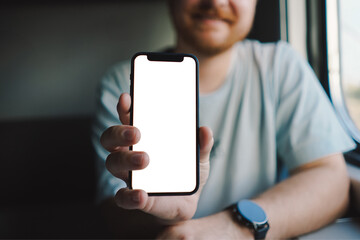 A man with a beard and mustache in a blue t-shirt is using a smartphone while traveling by Railway train, sitting in the train and looking out the window.