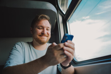 A man with a beard and mustache in a blue t-shirt is using a smartphone while traveling by Railway train, sitting in the train and looking out the window.