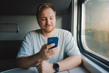 A man with a beard and mustache in a blue t-shirt is using a smartphone while traveling by Railway...