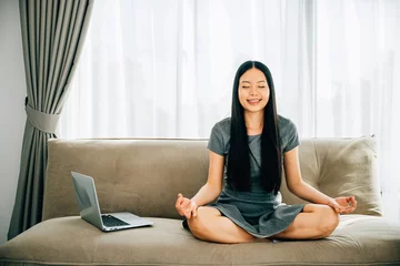 Tuinposter A serene Asian woman on sofa uses laptop meditating in lotus pose. Businesswoman finds relaxation and balance while practicing mindfulness online. Smiling student embodies harmony in the office. © sorapop