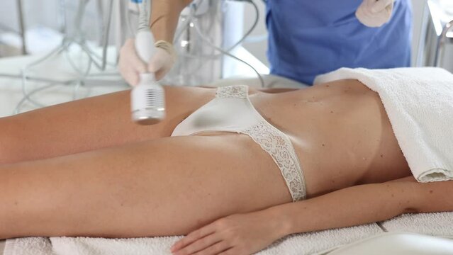 Aesthetician administering hardware procedure on female patient in cosmetology clinic, using ice hammer attachment to reduce body fat and contour thighs