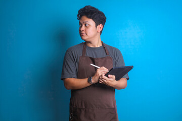 Young Asian barista man in brown apron work in coffee shop hold digital tablet on blue background