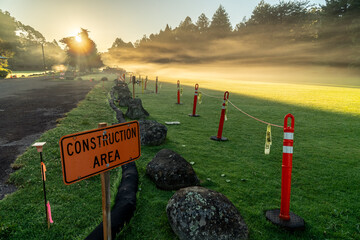 Orange plastic warning poles holding a safety line with "construction area" sign in bright color on a foggy grassy area basking in the morning sun, Kokee State Park, Kauai, Hawaii
