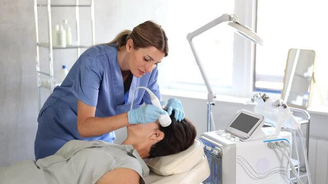 Woman doctor uses hardware ultrasonic to revitalize and tighten facial skin client at cosmetology clinic. High quality 4k footage