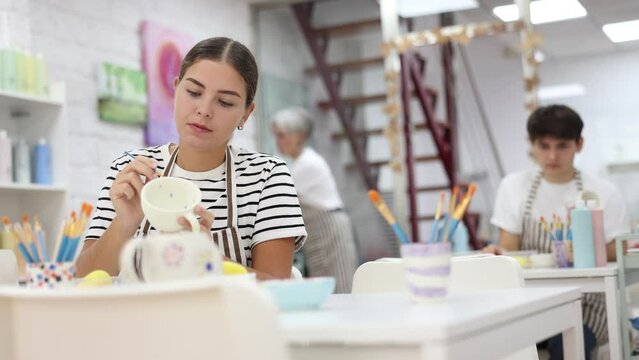 Cheerful young couple, guy and girl, actively engaged in creative hobby, decorating ceramic pottery together at table in cozy workshop setting