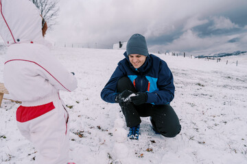 Fototapeta na wymiar Little girl looks at dad making a little snowman on a snowy hill