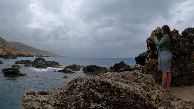 Shot of female tourist taking phots of waves crashing along the rocky beach in Malta on a cloudy day.