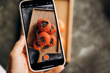 Woman taking photo of persimmon fruit with smartphone on grey background