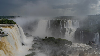 An impressive waterfall landscape. Streams of foaming water cascades into the abyss from the rocks. Tropical vegetation on ledges. Spray and fog rise into the sky. Poor visibility. Iguazu Falls.Brazil