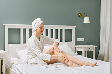 Young woman sitting on bed and massaging her legs using cream