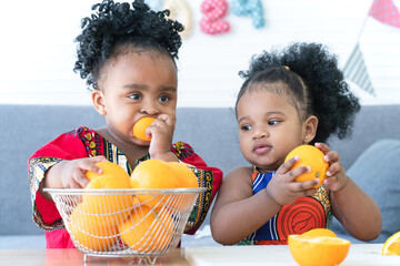 African cute kid girls enjoy eating fresh orange together at home. Two sister holding and squeeze orange in hands, eating orange juice from fresh orange. Healthy lifestyle