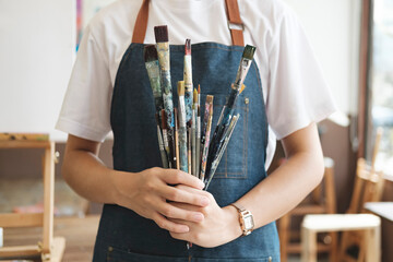 A close-up of the artist hands wearing an apron smeared with paint. Clutching many brushes and...