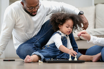 Cute little kid playing chess board toy as her parent sitting by. Multiracial family joyful game...