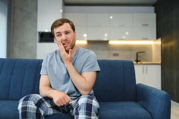 Frustrated young man touching his cheek while sitting on the couch at home