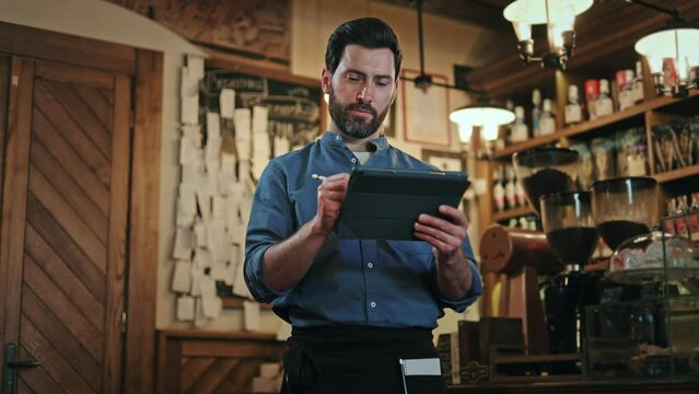 Serious man holding handheld order-taking device and inputting requests with stylus against backdrop of shelves with alcohol bottles. Concentrated cafe worker in uniform tending customers indoors.