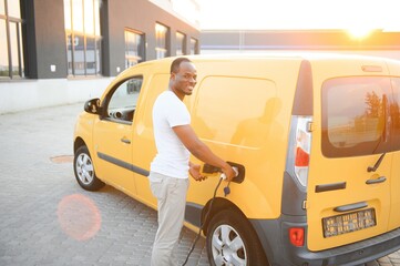 African american Man stands next to electric delivery vans at electric vehicle charging station