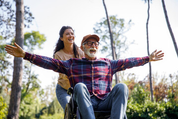 Social worker pushing cheerful retired senior man with arms outstretched in wheelchair at park