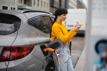Woman using smart phone and leaning on car at electric vehicle charging station