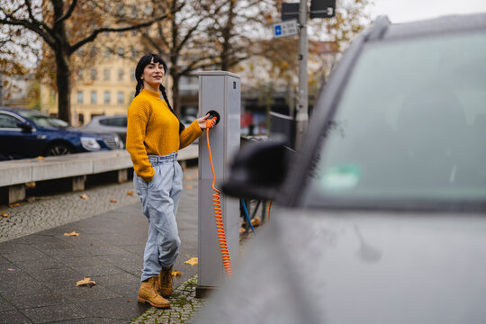 Smiling woman holding electric plug by car at charging station