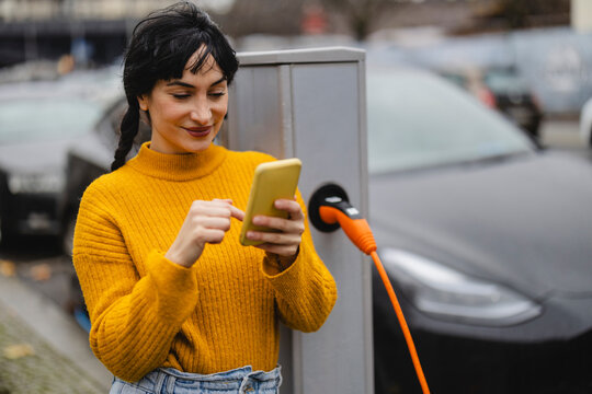 Smiling woman using smart phone at electric vehicle charging station