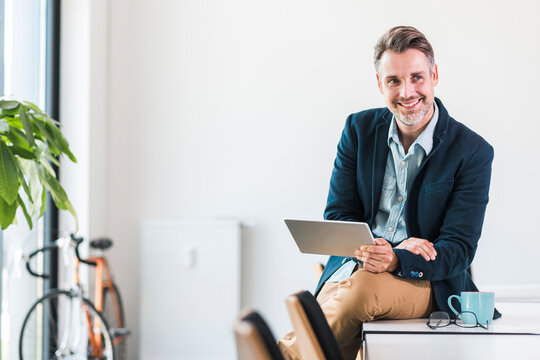 Smiling Businessman Holding Tablet PC And Sitting On Desk In Office